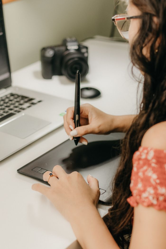 about-img Female graphic designer working with a tablet and laptop in a modern workspace.